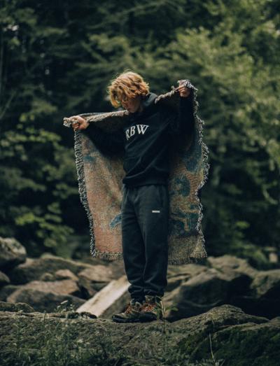 A man standing in nature wearing a Raised By Wolves sweatshirt and draping a blanket over his shoulders
