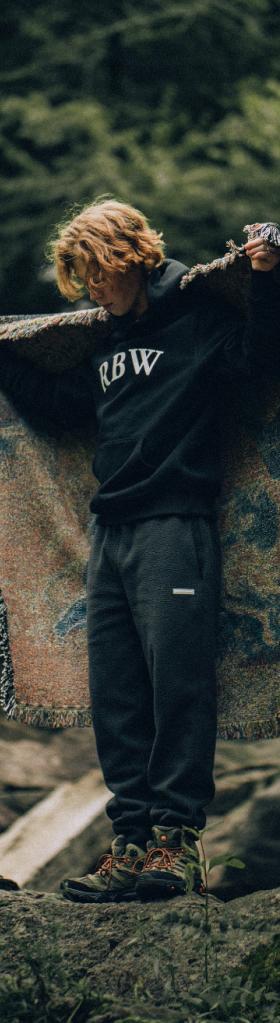 A man standing in nature wearing a Raised By Wolves sweatshirt and draping a blanket over his shoulders
