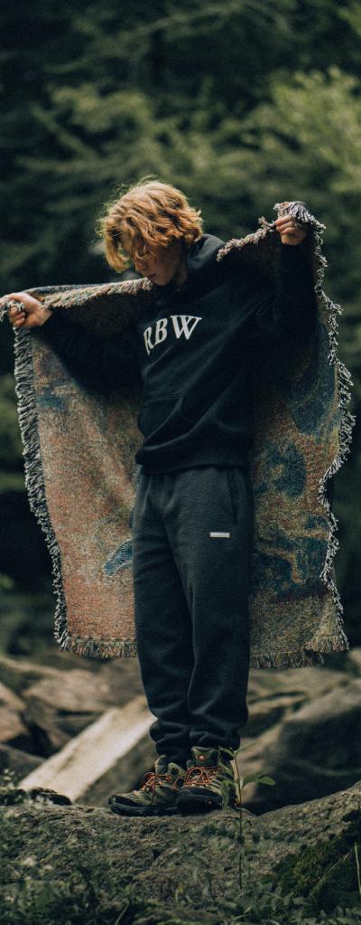 A man standing in nature wearing a Raised By Wolves sweatshirt and draping a blanket over his shoulders
