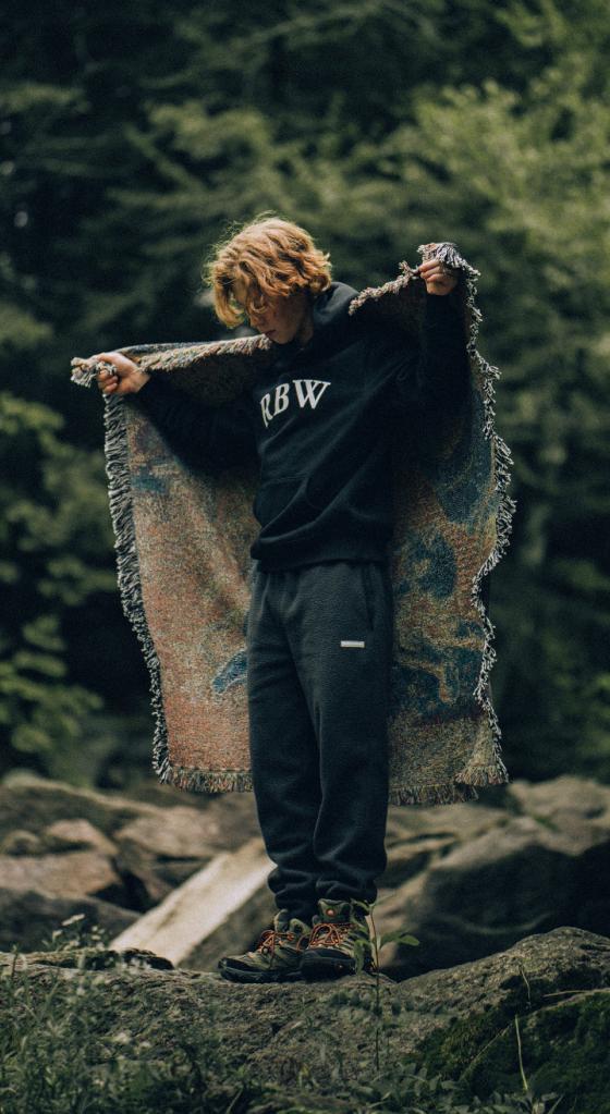 A man standing in nature wearing a Raised By Wolves sweatshirt and draping a blanket over his shoulders