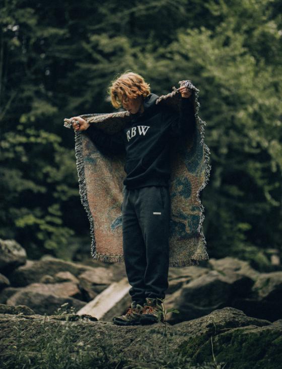 A man standing in nature wearing a Raised By Wolves sweatshirt and draping a blanket over his shoulders