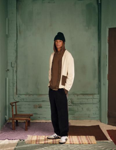 A man wearing a brown Kotn sweatshirt with a white cardigan over top of it along with a beanie, black pants and adidas shoes