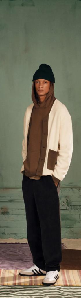 A man wearing a brown Kotn sweatshirt with a white cardigan over top of it along with a beanie, black pants and adidas shoes
