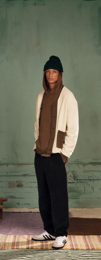 A man wearing a brown Kotn sweatshirt with a white cardigan over top of it along with a beanie, black pants and adidas shoes