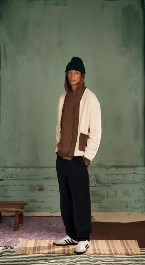A man wearing a brown Kotn sweatshirt with a white cardigan over top of it along with a beanie, black pants and adidas shoes