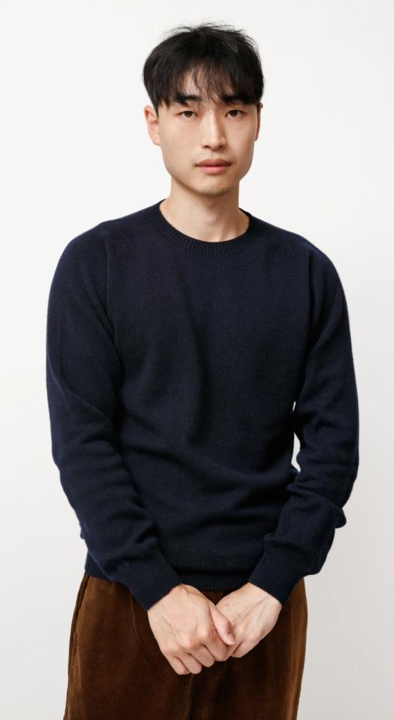 A man wearing a navy blue Neighbour sweatshirt and brown pants on a white backdrop