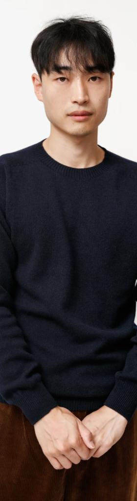 A man wearing a navy blue Neighbour sweatshirt and brown pants on a white backdrop