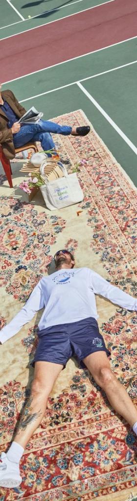 A man wearing a white Palmes Tennis long-sleeve while laying on a persian rug atop of tennis court