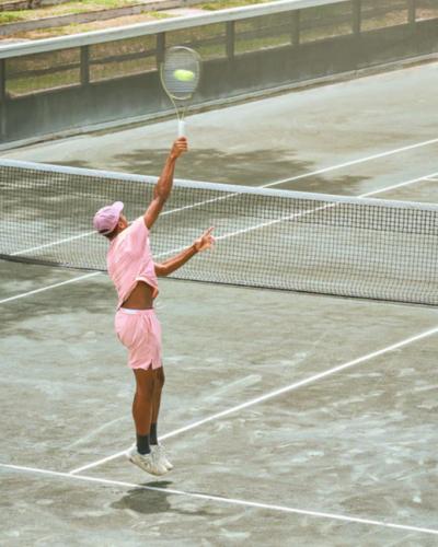 A man serving on a tennis court while wearing a pink Sigrun tennis outfit