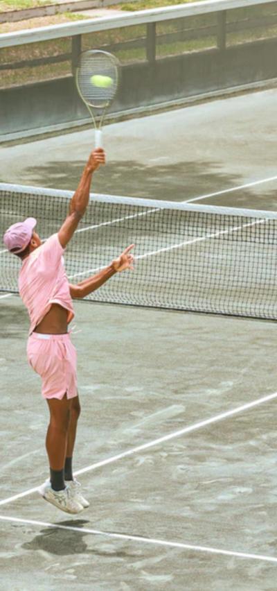 A man serving on a tennis court while wearing a pink Sigrun tennis outfit