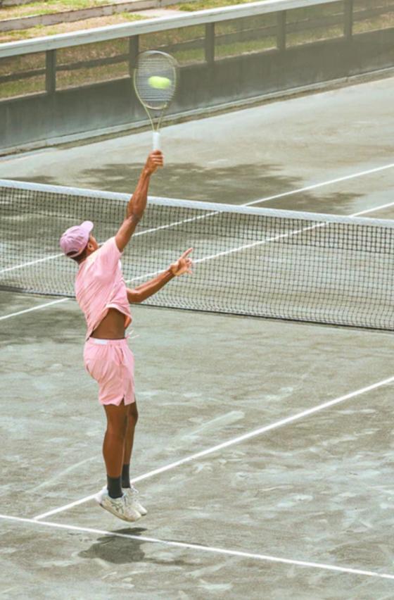 A man serving on a tennis court while wearing a pink Sigrun tennis outfit