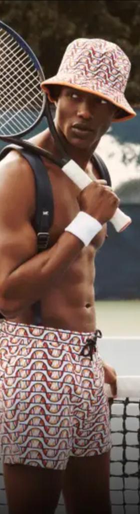 A man with a tennis racket over his shoulder wearing matching colorful Elesse shorts, backpack and bucket and no shirt
