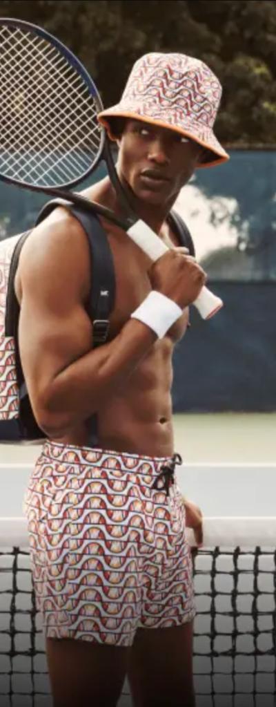 A man with a tennis racket over his shoulder wearing matching colorful Elesse shorts, backpack and bucket and no shirt