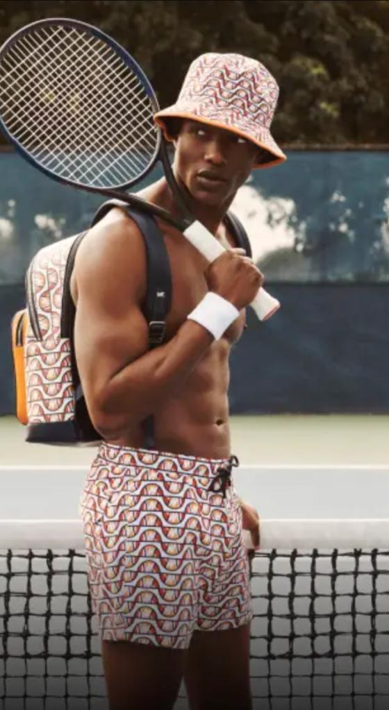 A man with a tennis racket over his shoulder wearing matching colorful Elesse shorts, backpack and bucket and no shirt