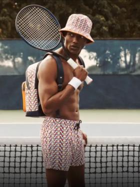 A man with a tennis racket over his shoulder wearing matching colorful Elesse shorts, backpack and bucket and no shirt