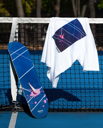 A white t-shirt with a tennis court print on it draped over a tennis court net with a matching skateboard balanced beside it