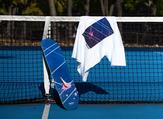 A white t-shirt with a tennis court print on it draped over a tennis court net with a matching skateboard balanced beside it