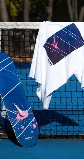 A white t-shirt with a tennis court print on it draped over a tennis court net with a matching skateboard balanced beside it