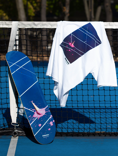 A white t-shirt with a tennis court print on it draped over a tennis court net with a matching skateboard balanced beside it