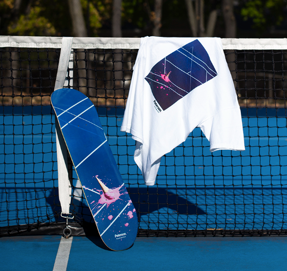 A white t-shirt with a tennis court print on it draped over a tennis court net with a matching skateboard balanced beside it