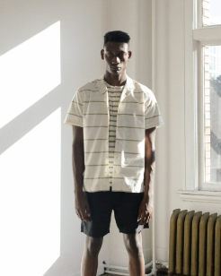 A man in a sunlit room wearing a Wings+Horns short sleeve striped button up and navy blue shorts