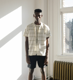 A man in a sunlit room wearing a Wings+Horns short sleeve striped button up and navy blue shorts