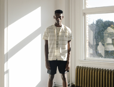 A man in a sunlit room wearing a Wings+Horns short sleeve striped button up and navy blue shorts