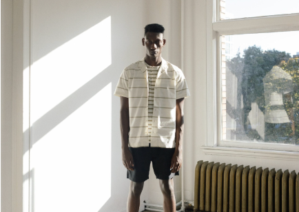 A man in a sunlit room wearing a Wings+Horns short sleeve striped button up and navy blue shorts