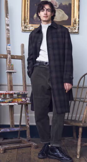A man wearing a brown checkered Oliver Spencer trench coat, a white turtleneck, green pants and black boots