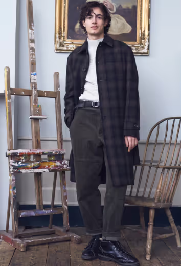 A man wearing a brown checkered Oliver Spencer trench coat, a white turtleneck, green pants and black boots