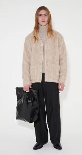 A man wearing a beige Stephan Schneider turtleneck and a beige cardigan over top along with black pants and bag
