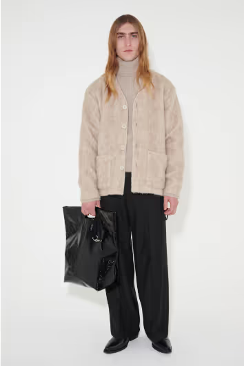 A man wearing a beige Stephan Schneider turtleneck and a beige cardigan over top along with black pants and bag