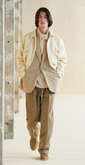 A man with long hair wearing a beige Auralee overshirt on top of a light brown blazer along with a turtle neck & brown pants