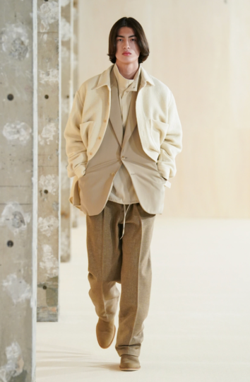 A man with long hair wearing a beige Auralee overshirt on top of a light brown blazer along with a turtle neck & brown pants