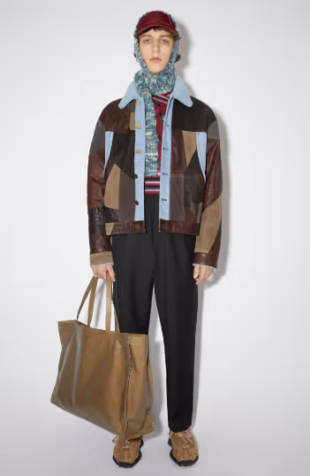 A man wearing an all over printed Acne Studios jacket with a colorful sweater underneath along with a scarf and brown bag