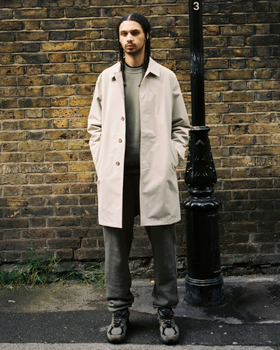 A man standing on the street wearing a biege JJJJound trench coat, green sweatshirt, dark grey pants and shoes