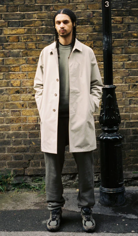 A man standing on the street wearing a biege JJJJound trench coat, green sweatshirt, dark grey pants and shoes
