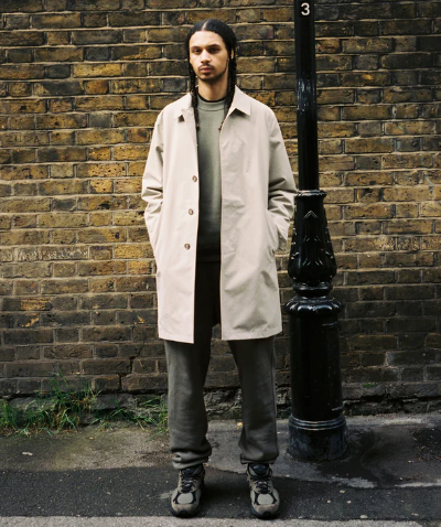 A man standing on the street wearing a biege JJJJound trench coat, green sweatshirt, dark grey pants and shoes