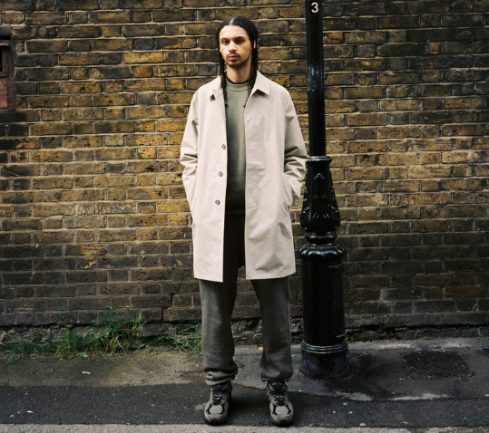A man standing on the street wearing a biege JJJJound trench coat, green sweatshirt, dark grey pants and shoes