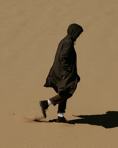 A man walking in the sand with a black Nanamica jacket wearing his hood with black pants, white socks and black boots on