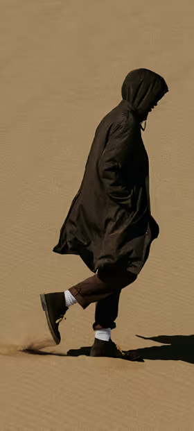 A man walking in the sand with a black Nanamica jacket wearing his hood with black pants, white socks and black boots on