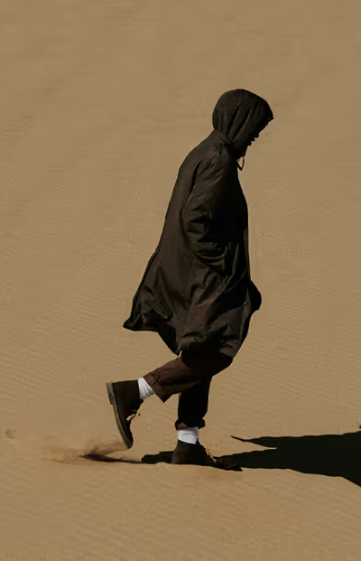 A man walking in the sand with a black Nanamica jacket wearing his hood with black pants, white socks and black boots on