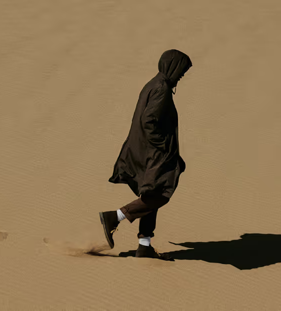 A man walking in the sand with a black Nanamica jacket wearing his hood with black pants, white socks and black boots on