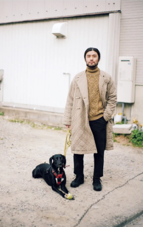 A man with a black dog wearing a beige Snow Peak jacket and a brown turtleneck along with black pants and shoes