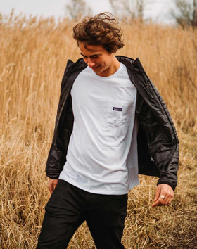 A man in a wheat field wearing a white Patagonia t-shirt along with black jacket and pants