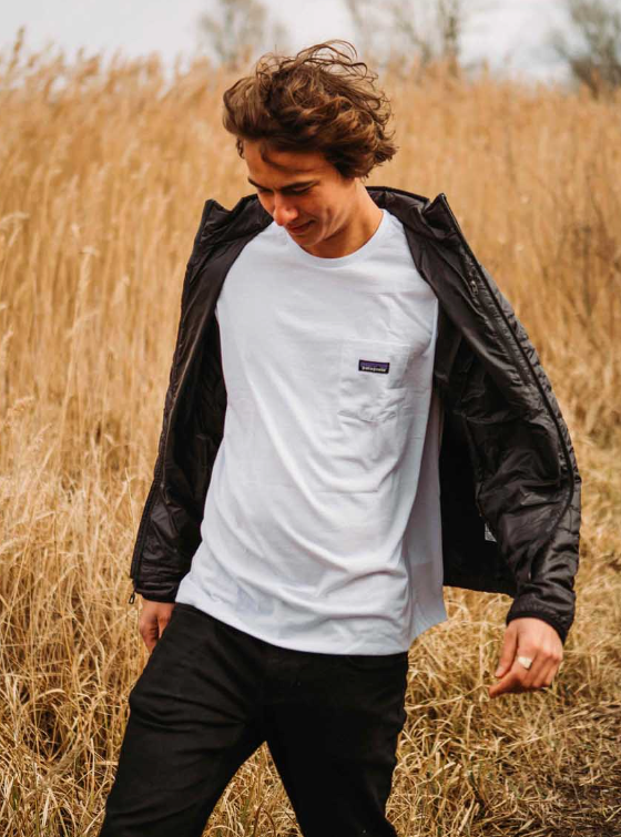A man in a wheat field wearing a white Patagonia t-shirt along with black jacket and pants