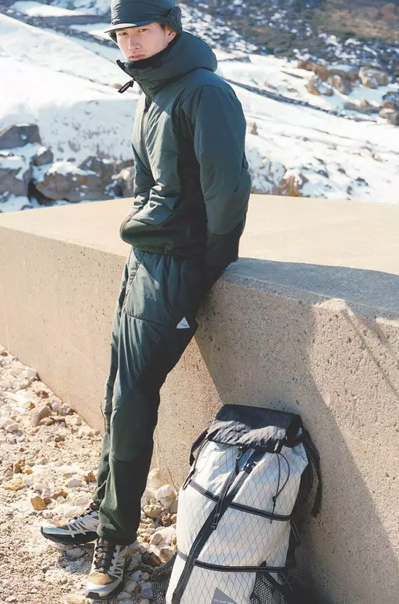 A man wearing a green And Wander snow suit with a matching green hat and salomon shoes