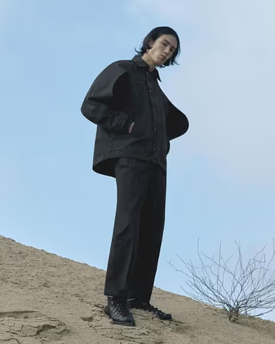 A man standing on a sandy hill wearing a black Konundrum jacket with matching black pants and boots