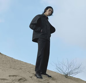 A man standing on a sandy hill wearing a black Konundrum jacket with matching black pants and boots