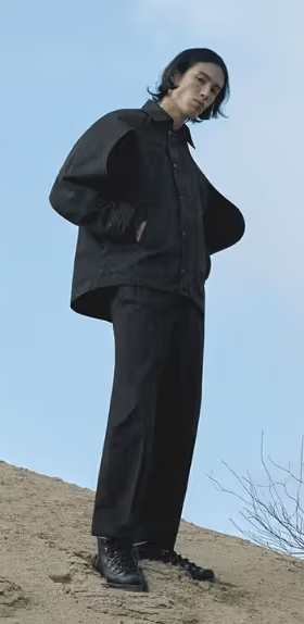 A man standing on a sandy hill wearing a black Konundrum jacket with matching black pants and boots